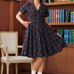 A woman models a short-sleeve plaid dress in a library setting with wooden bookshelves in the background. The dress features a navy blue, red, and green tartan pattern with a button-up front and a flared skirt.