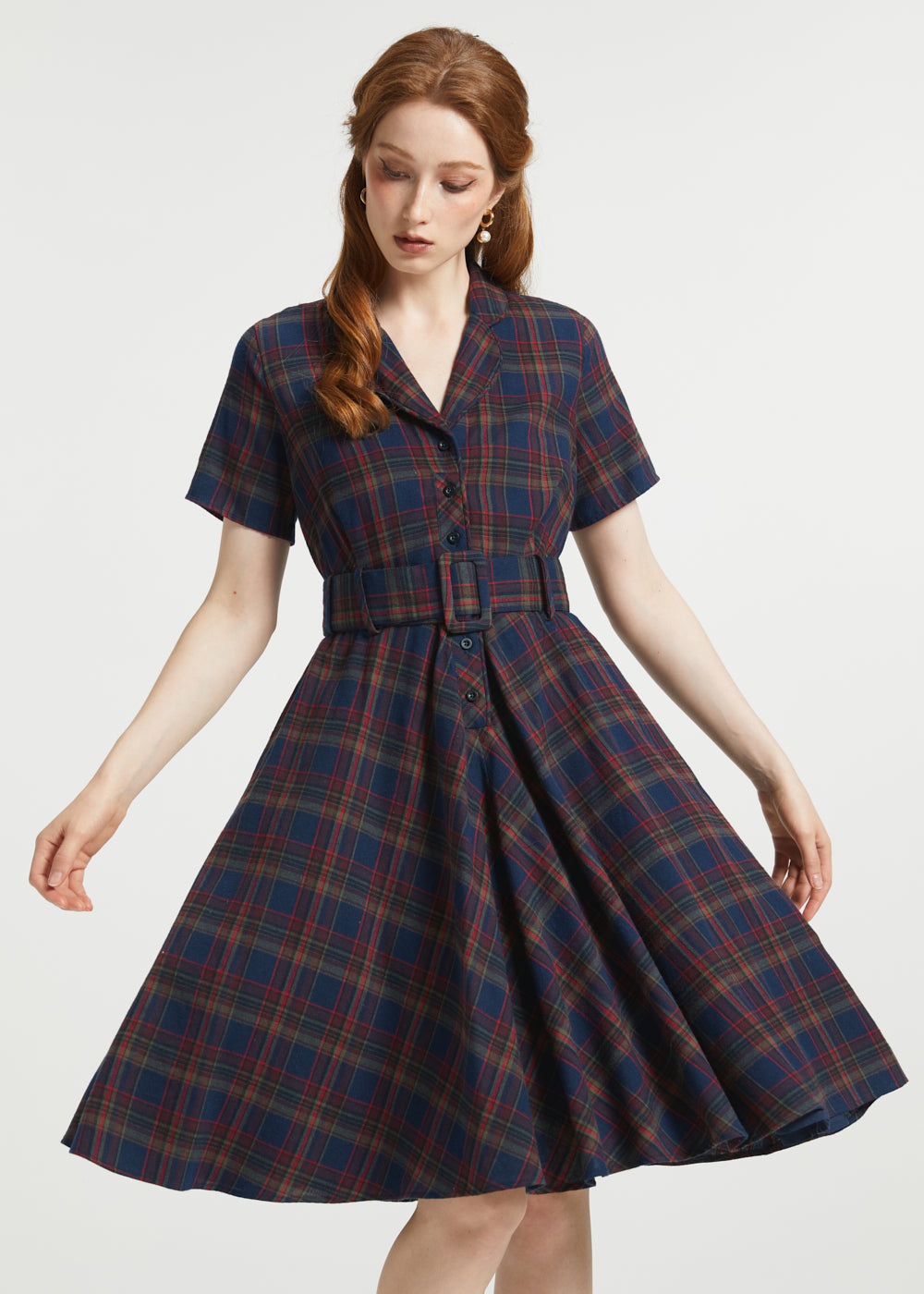 A woman modeling a short-sleeve plaid dress with a button front and matching belt, shown against a plain light gray studio background.