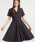 A woman modeling a short-sleeve plaid dress with a button front and matching belt, shown against a plain light gray studio background.
