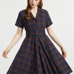 A woman modeling a short-sleeve plaid dress with a button front and matching belt, shown against a plain light gray studio background.