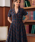 A woman wearing a short-sleeve plaid dress with a button front and matching belt, standing in front of a bookshelf.