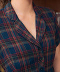 Close-up of a woman wearing a short-sleeve plaid shirt dress in navy blue with red, green, and beige tartan lines. The image shows the collar, chest, and upper torso against an indoor background.