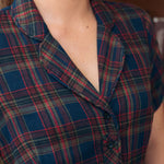 Close-up of a woman wearing a short-sleeve plaid shirt dress in navy blue with red, green, and beige tartan lines. The image shows the collar, chest, and upper torso against an indoor background.