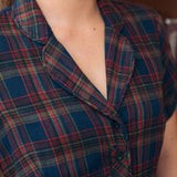 Close-up of a woman wearing a short-sleeve plaid shirt dress in navy blue with red, green, and beige tartan lines. The image shows the collar, chest, and upper torso against an indoor background.