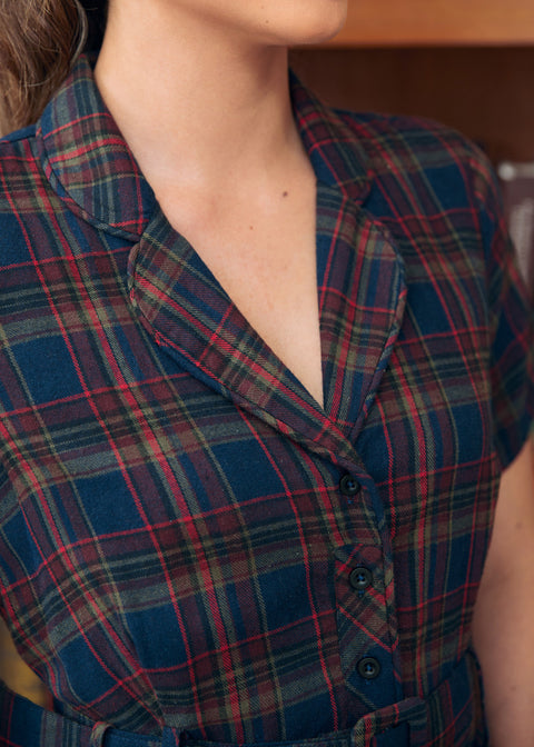 Close-up of a woman wearing a short-sleeve plaid shirt dress in navy blue with red, green, and beige tartan lines. The image shows the collar, chest, and upper torso against an indoor background.