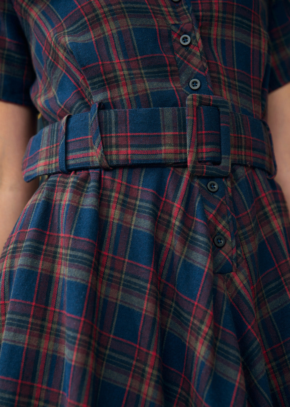 Close-up of a plaid dress with a matching belt, featuring blue, red, and green tartan pattern with dark buttons.