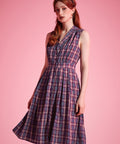 A woman modeling a sleeveless plaid dress with a V-neckline and gathered waist, standing against a solid pink studio background.