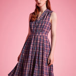 A woman modeling a sleeveless plaid dress with a V-neckline and gathered waist, standing against a solid pink studio background.