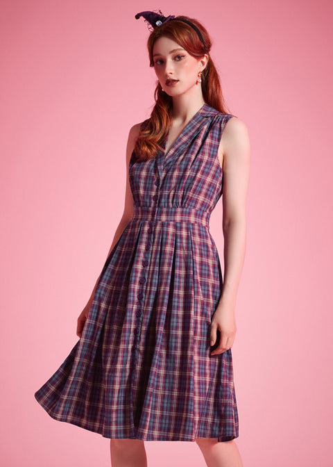 A woman modeling a sleeveless plaid dress with a V-neckline and gathered waist, standing against a solid pink studio background.