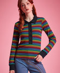 A woman modeling a long-sleeve knit polo shirt with horizontal rainbow stripes and a black collar, placket, and cuffs, paired with blue jeans against a solid pink background.