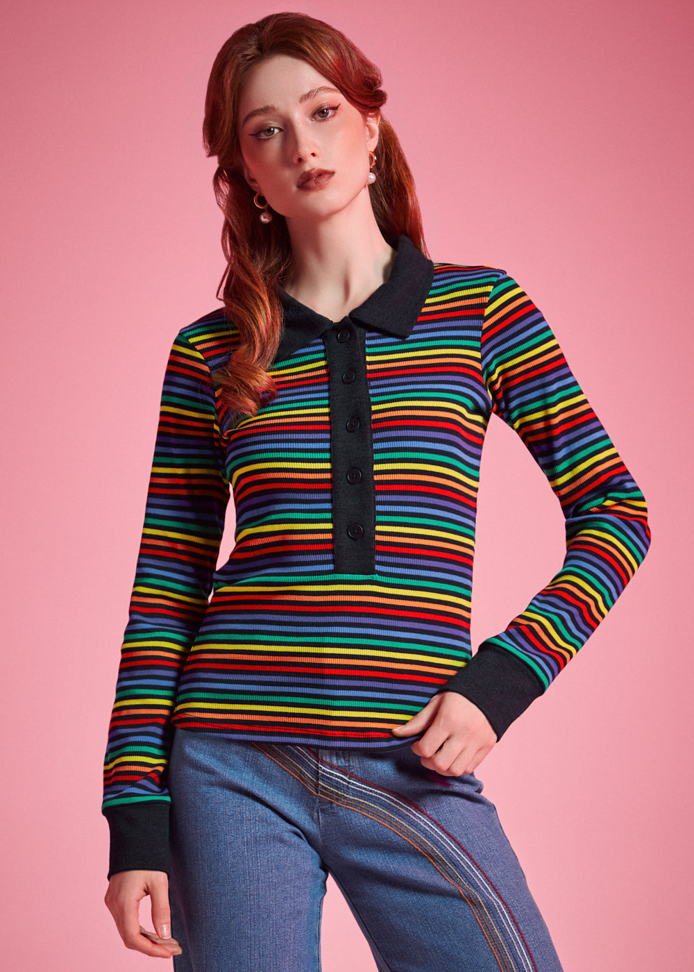 A woman modeling a long-sleeve knit polo shirt with horizontal rainbow stripes and a black collar, placket, and cuffs, paired with blue jeans against a solid pink background.