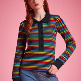 A woman modeling a long-sleeve knit polo shirt with horizontal rainbow stripes and a black collar, placket, and cuffs, paired with blue jeans against a solid pink background.