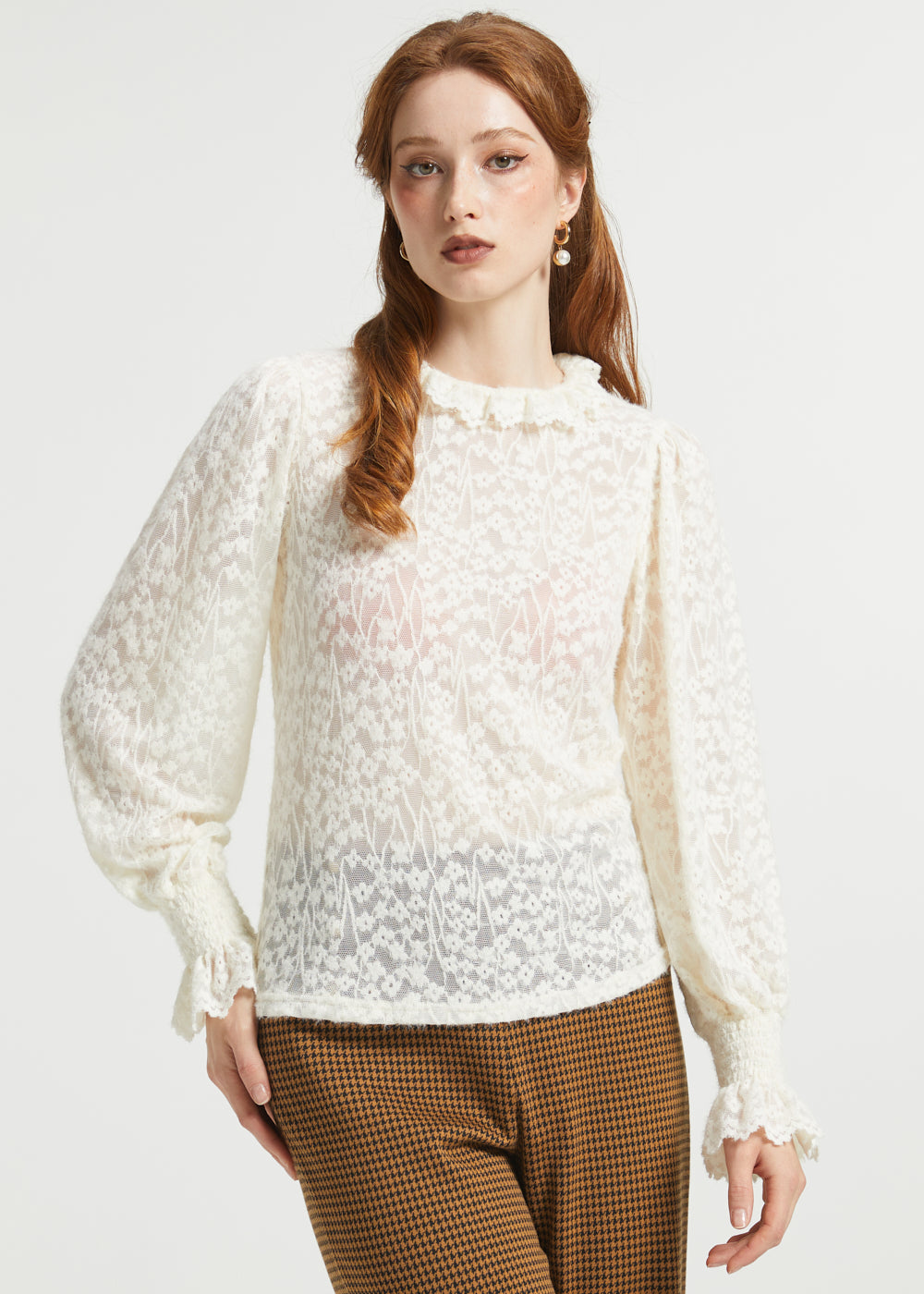 A woman modeling a cream-colored, semi-sheer lace long-sleeve blouse with a ruffled high neckline and gathered cuffs, paired with brown and black checkered trousers, against a plain light gray background.