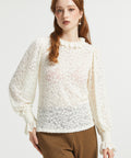A woman modeling a cream-colored, semi-sheer lace long-sleeve blouse with a ruffled high neckline and gathered cuffs, paired with brown and black checkered trousers, against a plain light gray background.