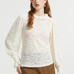 A woman modeling a cream-colored, semi-sheer lace long-sleeve blouse with a ruffled high neckline and gathered cuffs, paired with brown and black checkered trousers, against a plain light gray background.