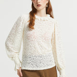 A woman modeling a cream-colored, semi-sheer lace long-sleeve blouse with a ruffled high neckline and gathered cuffs, paired with brown and black checkered trousers, against a plain light gray background.