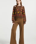 A woman stands against a light gray studio background wearing a brown knit sweater with colorful dinosaur motifs and matching brown striped wide-leg pants, paired with dark red heels.