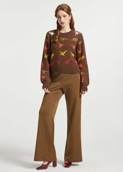 A woman stands against a light gray studio background wearing a brown knit sweater with colorful dinosaur motifs and matching brown striped wide-leg pants, paired with dark red heels.