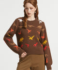 A woman modeling a brown crewneck sweater with a colorful dinosaur print, paired with brown and gold checkered trousers, against a light gray studio background.