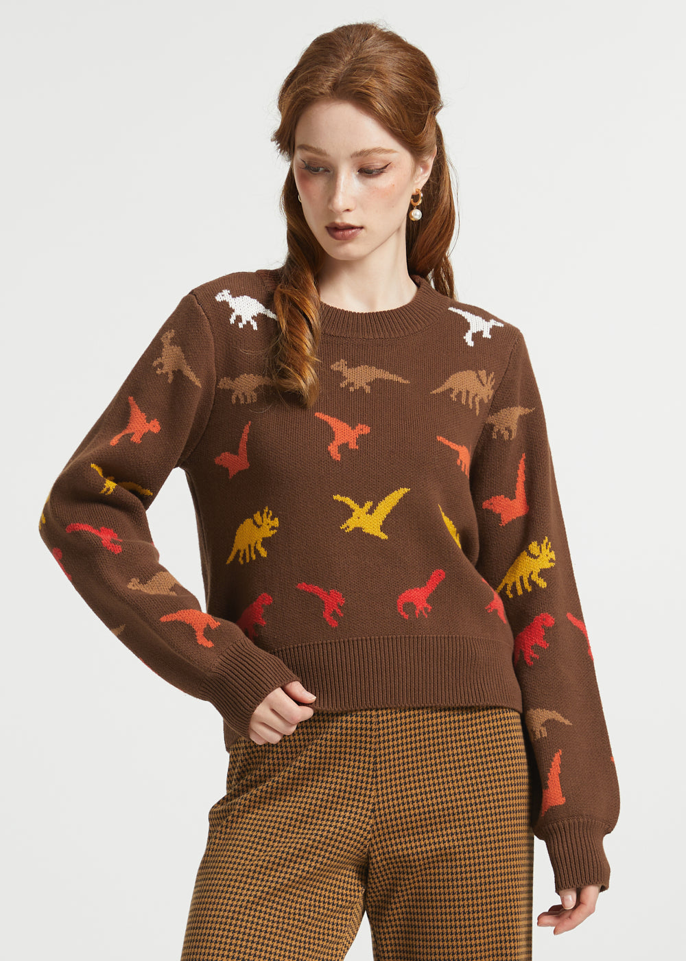 A woman modeling a brown crewneck sweater with a colorful dinosaur print, paired with brown and gold checkered trousers, against a light gray studio background.