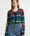 A woman modeling a dark navy long-sleeve button front cardigan with a colorful geometric and fruit knit pattern, paired with blue jeans, against a plain light gray studio background.