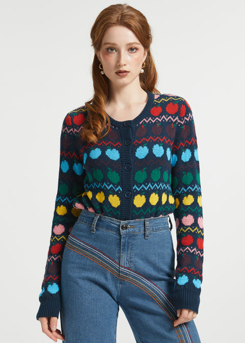 A woman modeling a dark navy long-sleeve button front cardigan with a colorful geometric and fruit knit pattern, paired with blue jeans, against a plain light gray studio background.