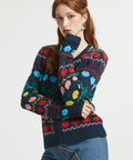 A woman wearing a navy blue knit cardigan with a colorful, multicolored geometric apple pattern, paired with blue jeans, against a plain light gray background.