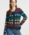 A woman modeling a dark navy blue button-up cardigan with a colorful Fair Isle pattern of red, yellow, green, blue, and pink apples, paired with blue jeans against a light gray studio background.