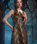 A woman modeling a sleeveless A-line dress with a dense multicolor floral print over a dark base, featuring a contrasting white collar, posed in front of a gothic-style arched window backdrop.