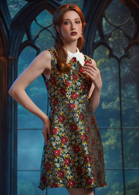 A woman modeling a sleeveless A-line dress with a dense multicolor floral print over a dark base, featuring a contrasting white collar, posed in front of a gothic-style arched window backdrop.