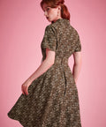 Back view of a woman modeling a short-sleeve fit-and-flare dress with a small floral print in olive green and cream, posed against a solid pink studio background.