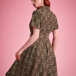 Back view of a woman modeling a short-sleeve fit-and-flare dress with a small floral print in olive green and cream, posed against a solid pink studio background.