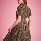 Back view of a woman modeling a short-sleeve fit-and-flare dress with a small floral print in olive green and cream, posed against a solid pink studio background.