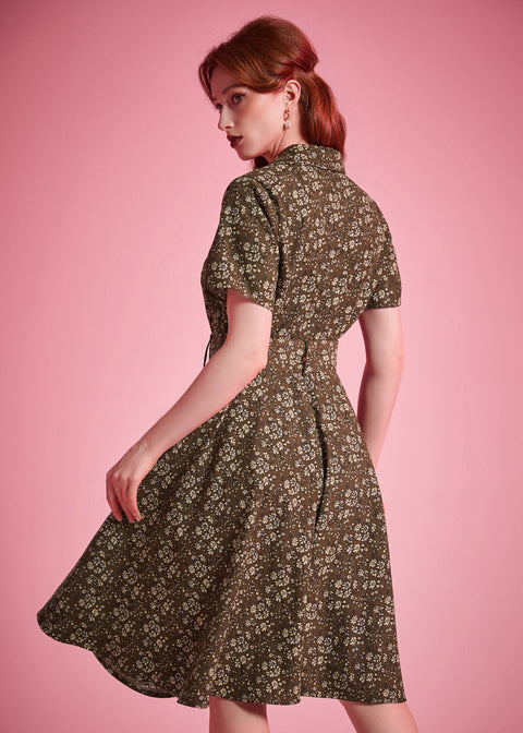 Back view of a woman modeling a short-sleeve fit-and-flare dress with a small floral print in olive green and cream, posed against a solid pink studio background.
