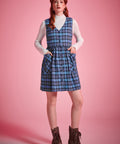 Full-length view woman wearing a blue plaid cotton jumper dress over a white mockneck top with brown lace-up ankle boots