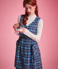 Woman wearing blue plaid V-neck jumper dress over a white mockneck top while holding playing cards