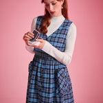 Woman wearing blue plaid V-neck jumper dress over a white mockneck top while holding playing cards