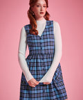 Woman wearing a blue plaid cotton jumper dress layered over a white mockneck top, showing V-neck and button-front design