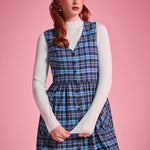 Woman wearing a blue plaid cotton jumper dress layered over a white mockneck top, showing V-neck and button-front design