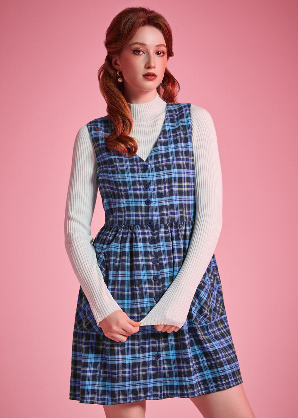Woman wearing a blue plaid cotton jumper dress layered over a white mockneck top, showing V-neck and button-front design