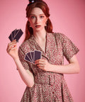 A woman with red hair wearing a small black witch hat and a short-sleeve floral dress, holding cards against a solid pink background.