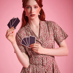 A woman with red hair wearing a small black witch hat and a short-sleeve floral dress, holding cards against a solid pink background.