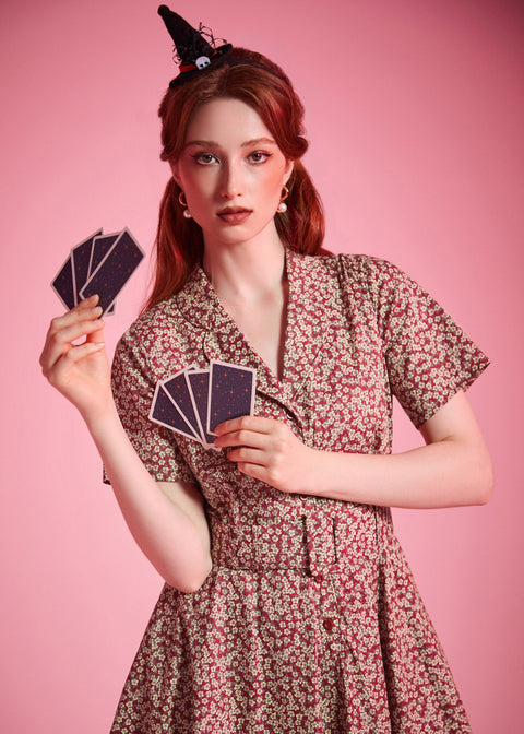 A woman with red hair wearing a small black witch hat and a short-sleeve floral dress, holding cards against a solid pink background.