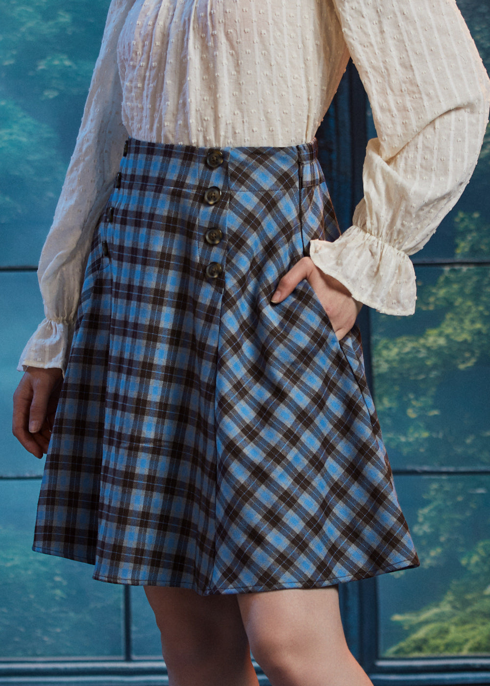 Three-quarter view of woman wearing blue brown plaid flare skirt with her hand in the side pocket, showing bias-cut side panel