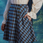 Three-quarter view of woman wearing blue brown plaid flare skirt with her hand in the side pocket, showing bias-cut side panel