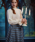 Woman wearing the ModCloth Sailor Flare blue plaid skirt with ivory high-neck blouse while holding a lit three-candle candelabra