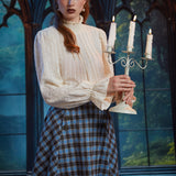 Woman wearing the ModCloth Sailor Flare blue plaid skirt with ivory high-neck blouse while holding a lit three-candle candelabra