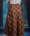 Back view of woman wearing a brown and tan plaid midi skirt showing sweeping A-line silhouette and braided belt
