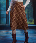 Woman wearing brown plaid A-line midi skirt with braided belt and hand in side pocket, styled with white blouse and boots