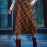 Woman wearing brown plaid A-line midi skirt with braided belt and hand in side pocket, styled with white blouse and boots
